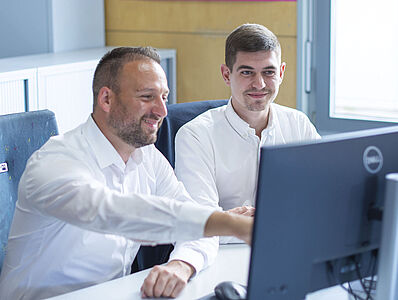 Two men are sitting at a desk in front of a monitor, with one man pointing at it.