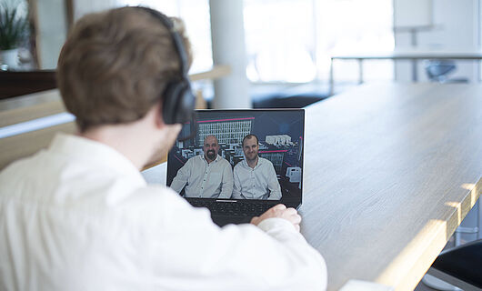 A man sits at a table in front of a laptop, watching a webinar featuring two presenters.
