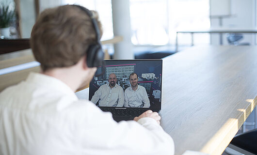 Ein Mann sitzt an einem Tisch vor einem Laptop und schaut sich ein Webinar mit zwei Sprechern an.
