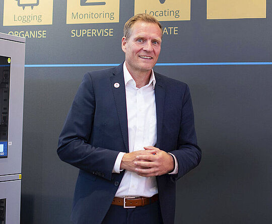 Lächelnder Mann steht vor einer Messewand neben Maschinen.