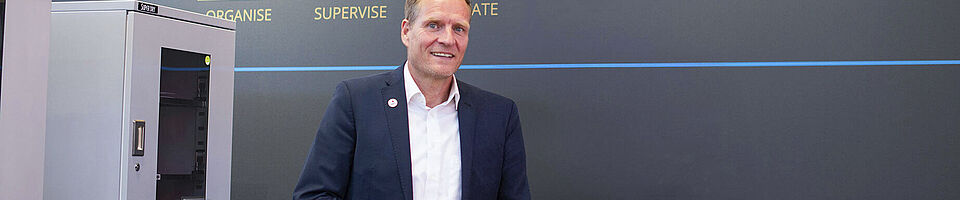 Smiling man stands in front of a trade fair wall next to machines.