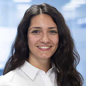 A woman wearing a white blouse stands smiling, looking toward the camera.