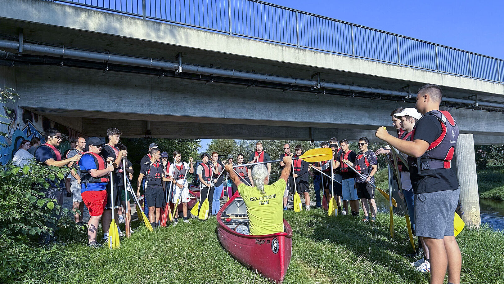 Viele junge Leute befinden sich unter einer Brücke am Ufer; in der Mitte sitzt ein Mann in einem Kanu.