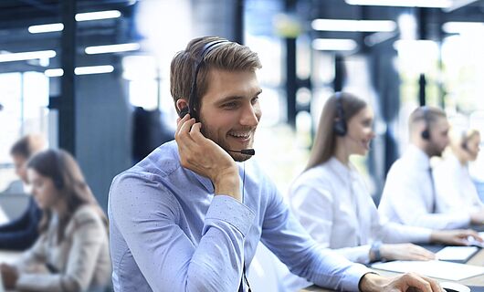 Mann mit Headset sitzt lächelnd in einem Callcenter am Schreibtisch, eine Hand am Ohr, die andere auf der Tastatur.
