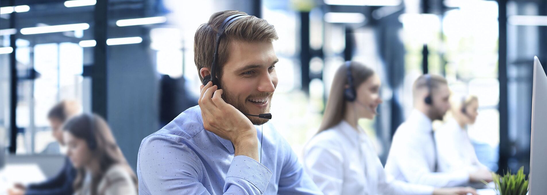 Mann mit Headset sitzt lächelnd in einem Callcenter am Schreibtisch, eine Hand am Ohr, die andere auf der Tastatur.