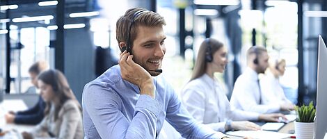 Smiling man wearing a headset sits at a desk in a call center, one hand on his ear, the other on the keyboard.