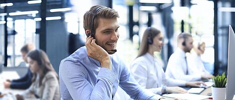 Smiling man wearing a headset sits at a desk in a call center, one hand on his ear, the other on the keyboard.