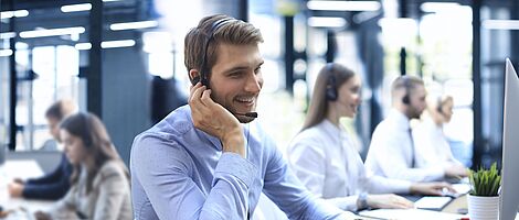 Mann mit Headset sitzt lächelnd in einem Callcenter am Schreibtisch, eine Hand am Ohr, die andere auf der Tastatur.