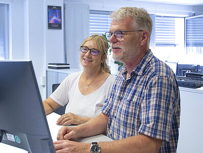 A man and a woman are standing at a desk in front of a monitor.
