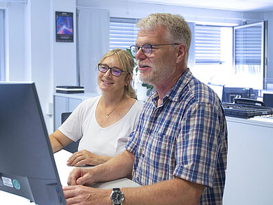 Ein Mann und eine Frau stehen an einem Schreibtisch vor einem Monitor.