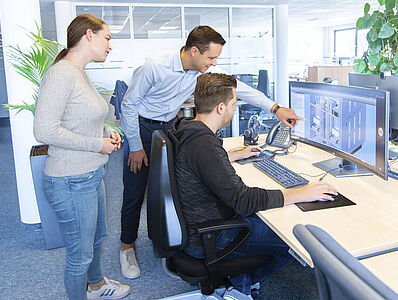 A man is sitting at a desk in an office working on a PC, another man is standing next to him pointing at the screen, and a woman is standing behind them