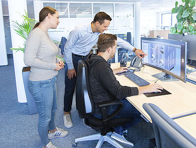 Ein Mann sitzt in einem Büro an einem Schreibtisch am PC, neben ihm steht ein Mann der auf den Bildschirm zeigt, Hinter ihm steht eine Frau.