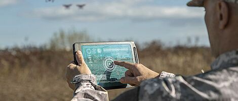 Military analysts oversee the coordination of the military team using drones. A military commander uses a digital tablet to direct augmented reality troops in the field.
