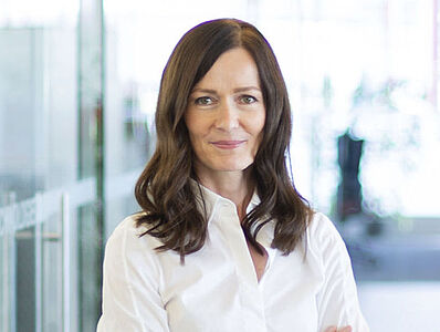 A woman wearing a white blouse stands smiling, looking toward the camera.
