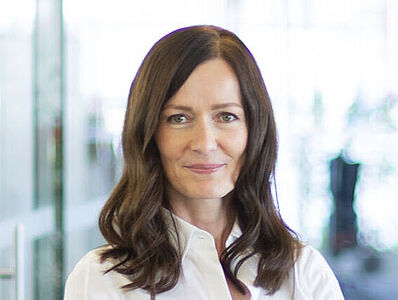 A woman wearing a white blouse stands smiling, looking toward the camera.