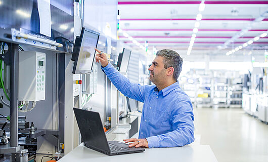 Man operates a machine via the control panel, with a standing desk and laptop next to him.