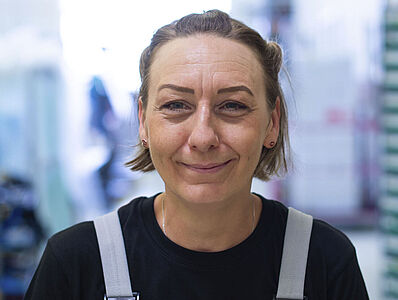 A smiling woman wearing overalls stands in a manufacturing hall.