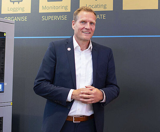 Smiling man stands in front of a trade fair wall next to machines.