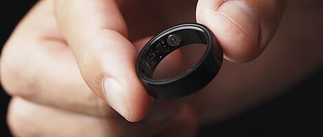 Eine Hand hält zwischen zwei Fingern einen schwarzen Smart Ring.