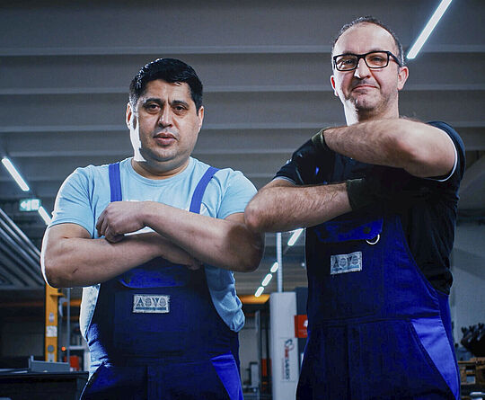Two men in workwear stand side by side in a manufacturing hall with their arms crossed.