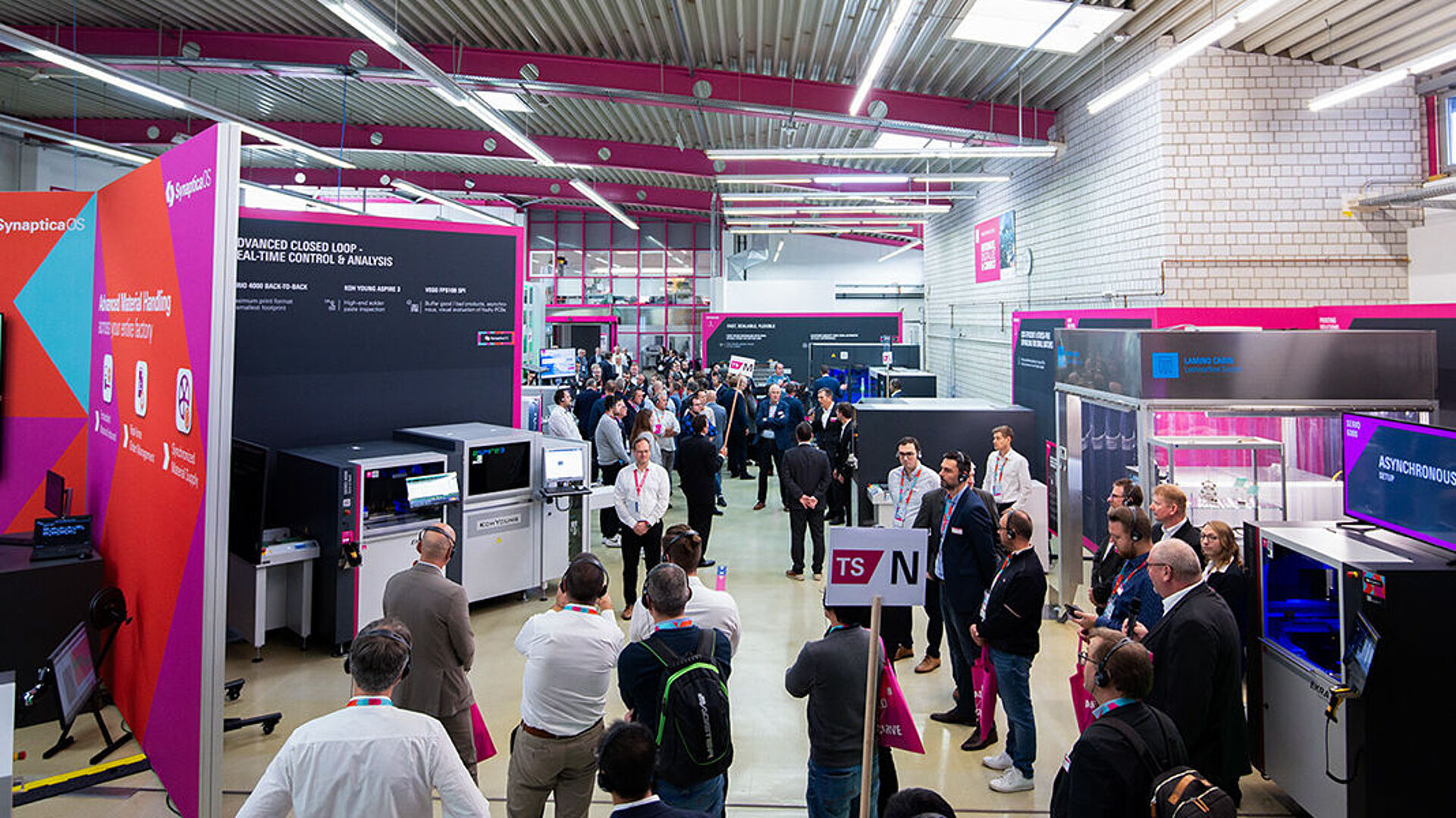 An interior view of a production hall with exhibition booths, machinery, and several people.