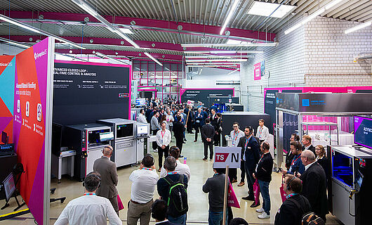 An interior view of a production hall with exhibition booths, machinery, and several people.