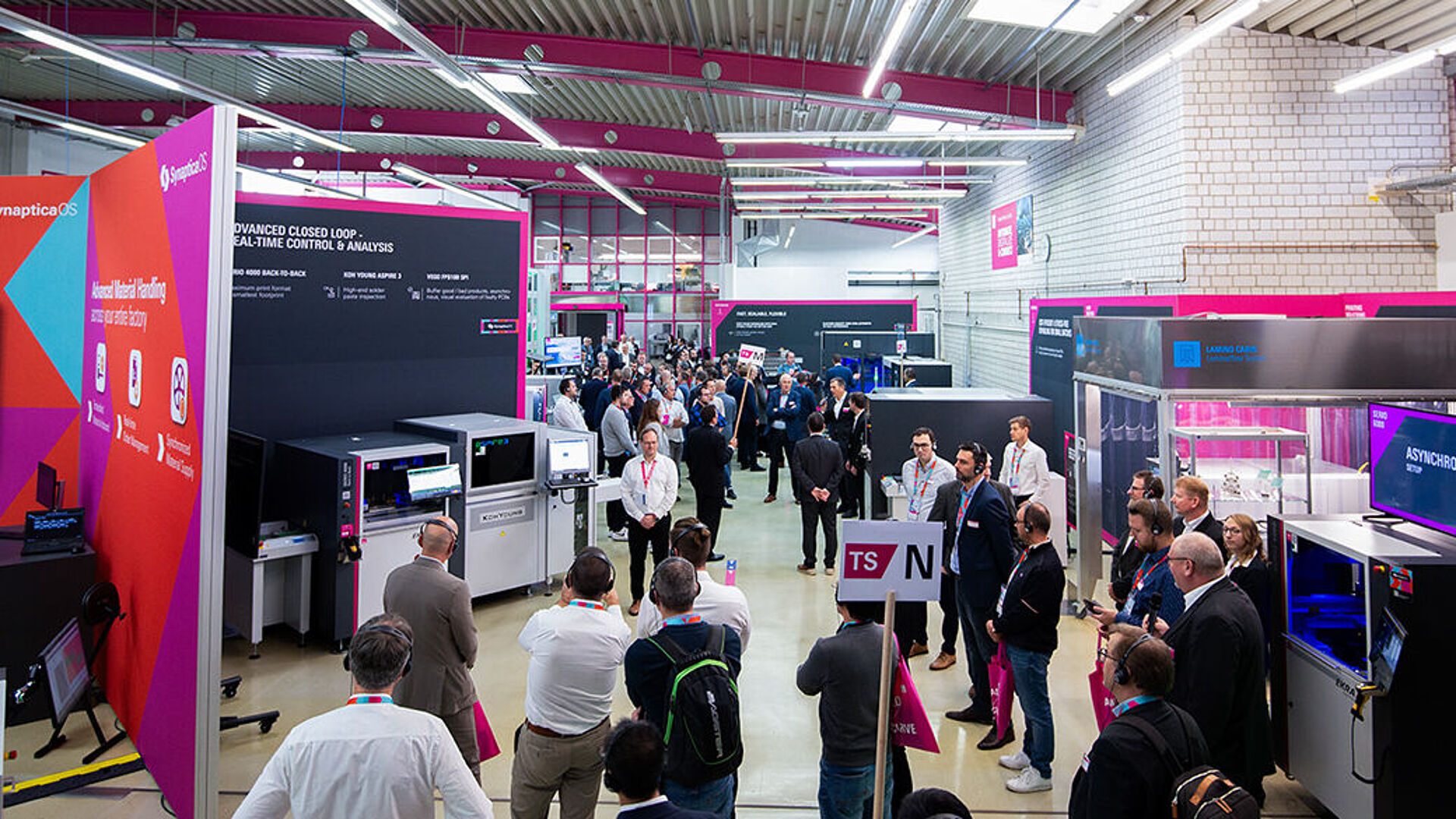 An interior view of a production hall with exhibition booths, machinery, and several people.