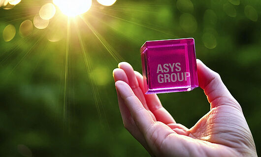 Magenta-colored cube with ASYS Group lettering is held in a hand towards the sunlight.