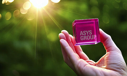 Magenta-colored cube with ASYS Group lettering is held in a hand towards the sunlight.