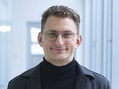 Smiling man in a black turtleneck sweater in a well-lit hallway.