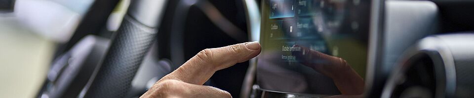 Innenraum eines modernen Autos: Eine Hand liegt am Lenkrad, die andere bedient den zentralen Touchscreen.