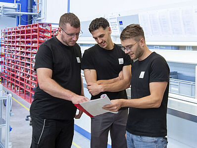 Drei Männer stehen in einer Produktionshalle und schauen gemeinsam auf ein Notizheft