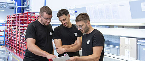 Drei Männer stehen in einer Produktionshalle und schauen gemeinsam auf ein Notizheft