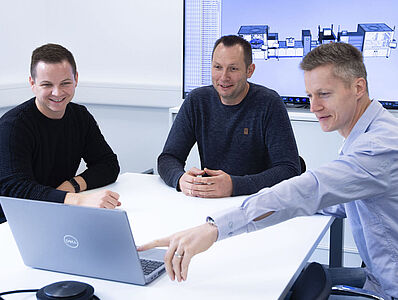 Three men are sitting at a table in a meeting room looking at a laptop, one of them pointing at the screen. There is a monitor in the background.