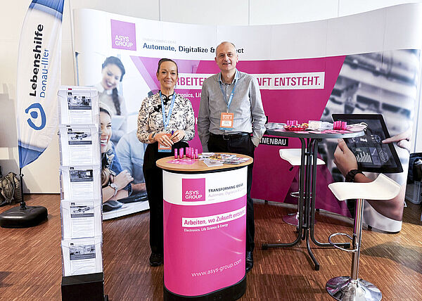 Ein Mann und eine Frau stehen nebeneinander auf einem Messestand hinter einem Stehtisch.