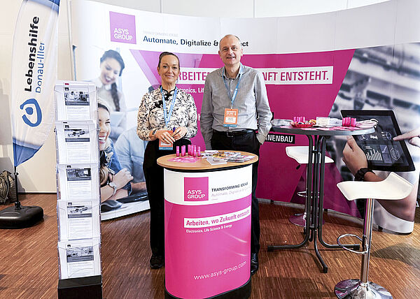 Ein Mann und eine Frau stehen nebeneinander auf einem Messestand hinter einem Stehtisch.