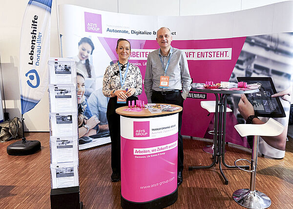 Ein Mann und eine Frau stehen nebeneinander auf einem Messestand hinter einem Stehtisch.