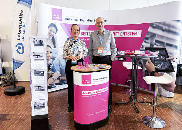 Ein Mann und eine Frau stehen nebeneinander auf einem Messestand hinter einem Stehtisch.