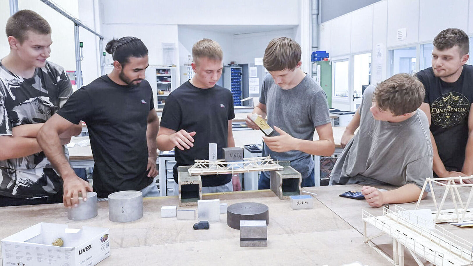 Six boys are standing at a workbench, testing the stability of a wooden bridge.