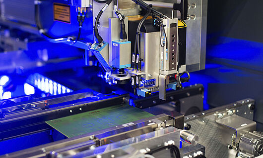 Interior view of a laser machine with an inserted circuit board. Blue light visible in the background.