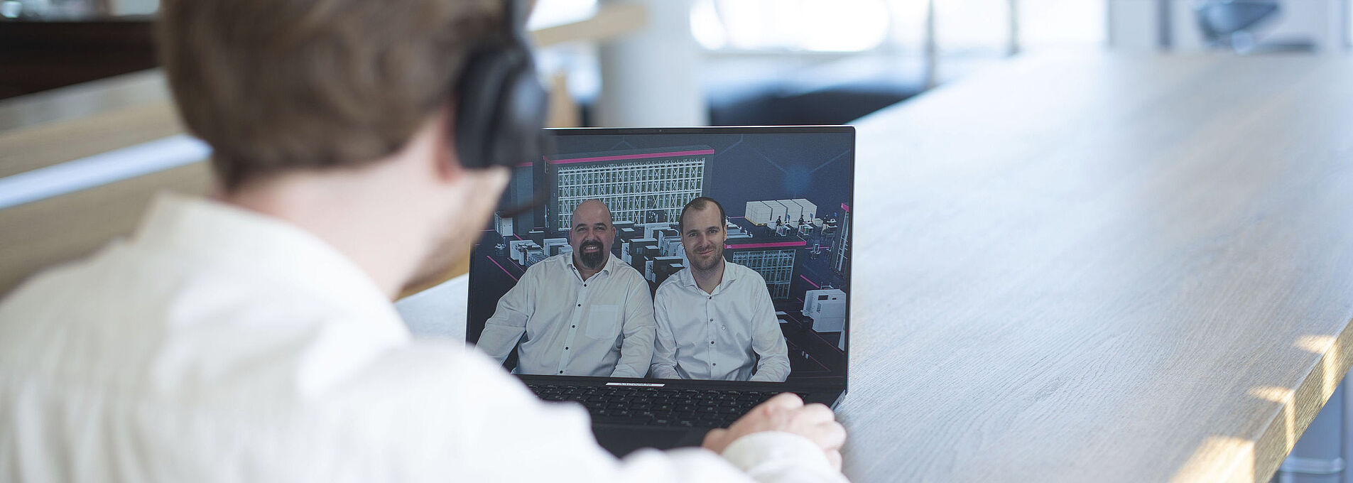 A man sits at a table in front of a laptop, watching a webinar featuring two presenters.