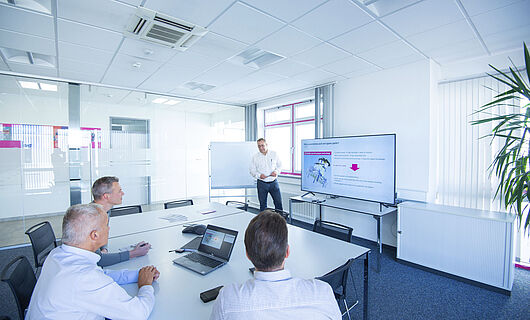 Three men are looking at a large monitor in a meeting room. One man is standing next to the monitor and giving a presentation.