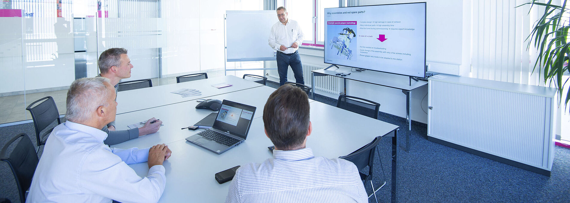 Drei Männer schauen auf einen großen Monitor in einem Besprechungszimmer. Ein Mann steht neben dem Monitor und präsentiert.