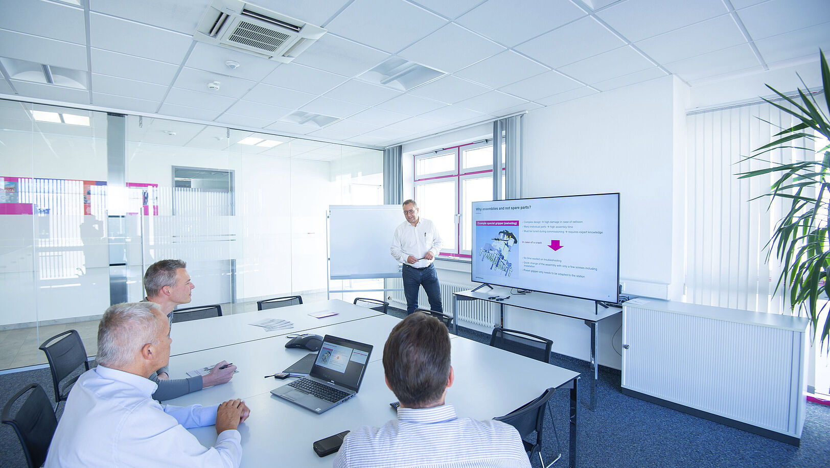 Three men are looking at a large monitor in a meeting room. One man is standing next to the monitor and giving a presentation.