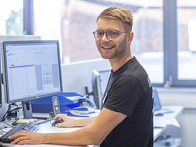 Ein junger Mann steht an einem Schreibtisch in einem Büro und hat die Hände auf Maus und Tastatur.