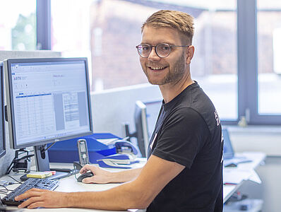 Young man standing at a desk in an office with his hands on a mouse and keyboard.