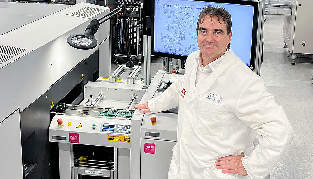 A man wearing a lab coat stands with his arm bent in front of a handling machine. In the background, more machines can be seen in a hall.