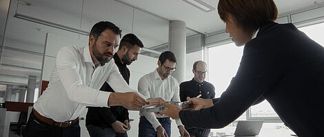 A man hands an object to a woman across the table. In the background, three other men are standing next to him.