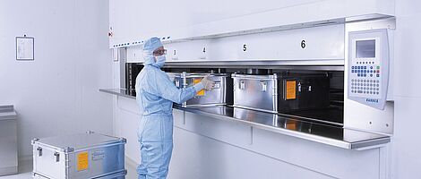 Man in full laboratory clothing at a cleanroom lift system with several metal containers.