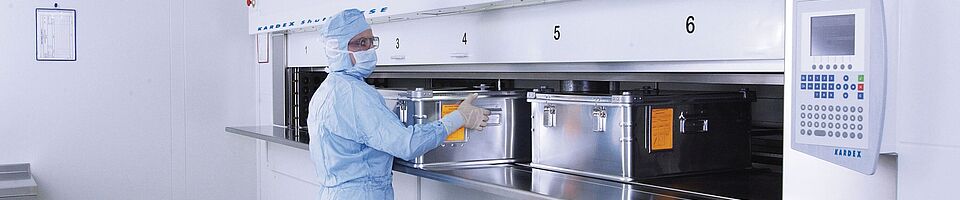 Mann in voller Laborkleidung an einem Reinraum-Liftsystem mit mehreren Metallkisten.